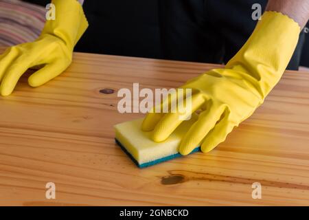 Mani dell'uomo con guanti gialli che puliscono un tavolo con una spugna Foto Stock