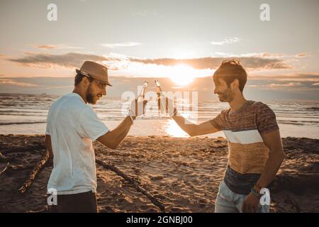 Giovani allegri che trascorrono del tempo insieme sulla spiaggia e bevono birra e tostano in spiaggia in vacanza al tramonto estivo al crepuscolo Foto Stock