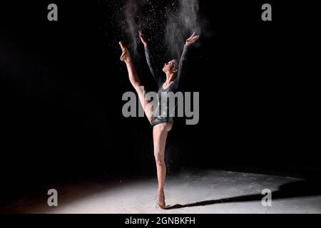 Ballerina flessibile donna danzante e spolverare la farina su sfondo nero studio, indossando tuta nera e scarpe a punta. Ritratto completo di attra Foto Stock