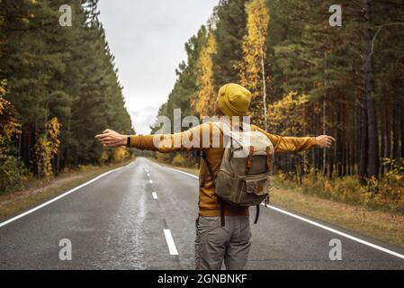 L'uomo in un cappello giallo e un maglione su una strada asfaltata liscia e vuota lungo una bella foresta dorata sta in piedi con le braccia distese ai lati. Foto Stock