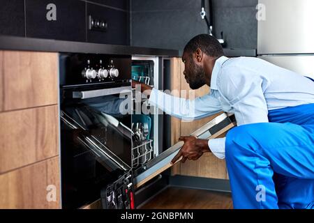 Il giovane professionista africano americano Handyman in tute blu uniforme sta per riparare la lavastoviglie, fiducioso appaltatore che la esamina prima della riparazione, Foto Stock