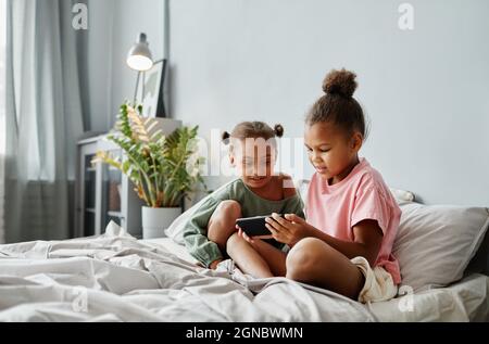 Ritratto di due ragazze afroamericane usando lo smartphone insieme a letto in una stanza accogliente, spazio copia Foto Stock