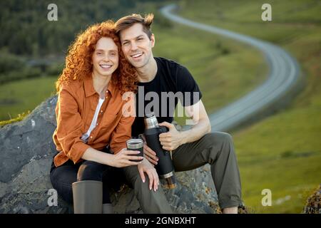 positivo giovane coppia caucasica siede prendendo pausa, avendo riposo sulle montagne, bevendo tè da thermos. Uomo rilassato e rosso ricci donna in casual w Foto Stock