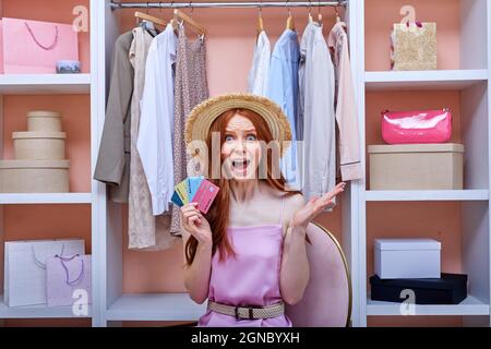 donna redhead ottimista tra molti vestiti nel suo armadio con smartphone e carta di credito banca di shopping online via internet, shopaholic ama shoppin Foto Stock