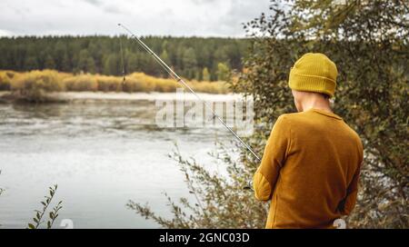 Il giovane pescatore in un maglione giallo e cappello sta pescando sulla riva del fiume nella foresta d'autunno con un verga che gira nelle sue mani. Concetto di relaxin Foto Stock