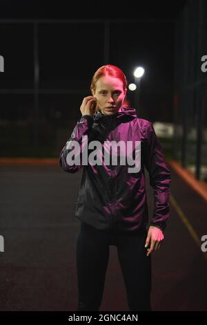 Ritratto di sicura donna caucasica con Ponytail indossando cuffie, guardando la Camera in posa. Giovane donna in Black Sportive Wear di notte in Cit Foto Stock