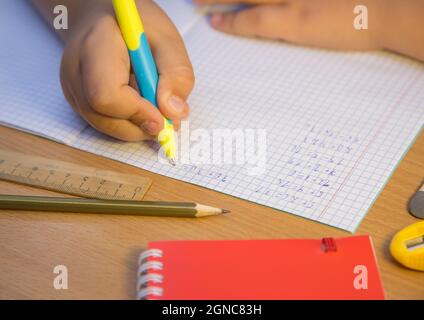 Il primo piano della mano della pupilla risolve gli esempi matematici con una penna a sfera. Uno scolaro svolge un compito sul posto di lavoro. Il concetto di educazione dei bambini, di insegnamento delle conoscenze, delle competenze e delle abilità. Foto Stock