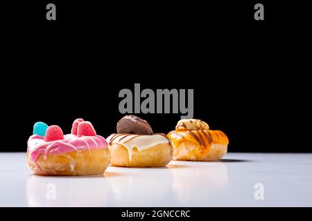 Fotografia di una composizione di tre ciambelle decorate con cioccolato colorato, fagioli di gelatina e biscotti di cioccolato. La foto è scattata su un tavolo bianco AN Foto Stock