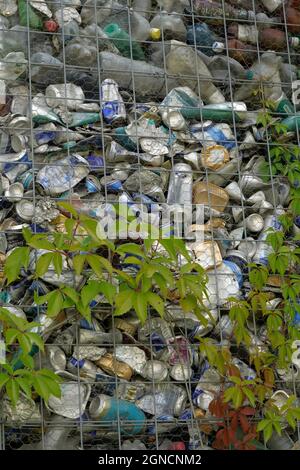 Dettaglio di bottiglie di plastica e lattine di alluminio schiacciate e compresse. Sono messi in una struttura metallica per recinzione. Ci sono piante striscianti che crescono. Foto Stock