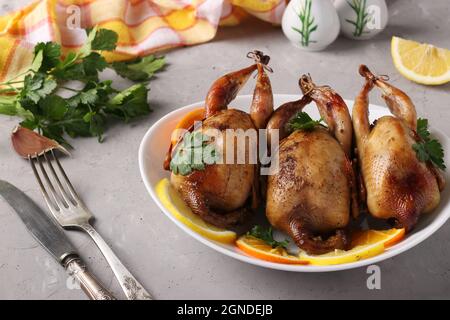 Tre quaglie al forno servite con limone, arancia e prezzemolo su un piatto bianco su sfondo grigio. Foto Stock