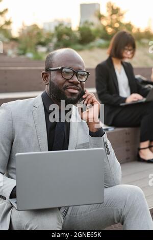 Ritratto verticale medio di bell'uomo afroamericano portatore di abito elegante seduta su panca all'aperto con computer portatile che parla al telefono con col Foto Stock