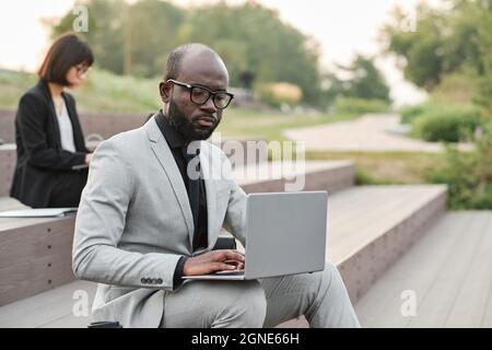 Ritratto orizzontale medio di grave uomo afroamericano maturo con barba sul viso indossando un elegante abito seduto in un parco urbano utilizzando un computer portatile Foto Stock