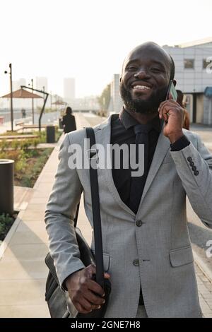 Verticale medio ritratto di gioioso bell'uomo afroamericano che cammina lungo la strada dopo aver finito il lavoro in ufficio avendo chiamata telefonica Foto Stock