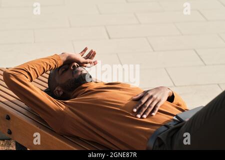 Orizzontale alto angolo di ripresa del giovane afro americano stanco rilassarsi su panca di legno all'aperto nella soleggiata giornata estiva Foto Stock