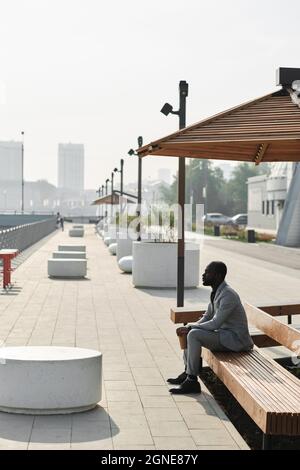 Foto lunga verticale del manager africano americano maturo indossando tuta grigia seduta sul terrapieno guardando il fiume pensando a qualcosa, spazio copia Foto Stock