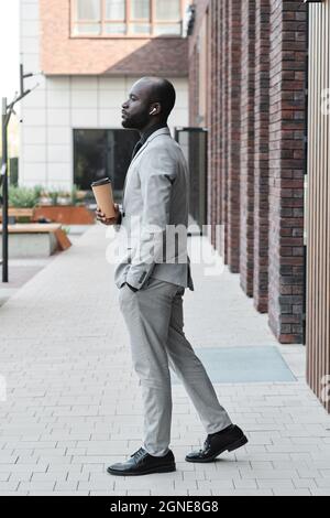 Scatto verticale a tutta lunghezza di un uomo afro-americano che indossa un elegante abito grigio in piedi all'aperto per ascoltare la musica in auricolari e drin Foto Stock