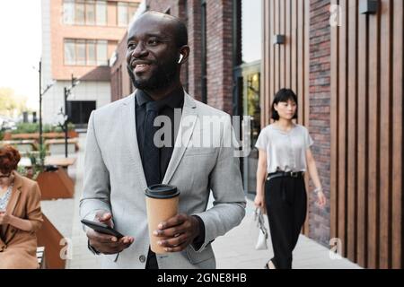 Ritratto di uomo afro-americano sicuro indossando elegante abito grigio in piedi all'aperto ascoltare musica in auricolari, bere caffè e chat Foto Stock