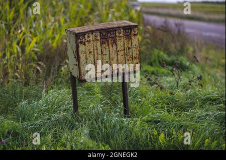 una vecchia cassetta postale gialla arrugginita da un campo di mais in una giornata di sole e una lunga erba che ondeggiano nel vento sotto la cassetta postale Foto Stock