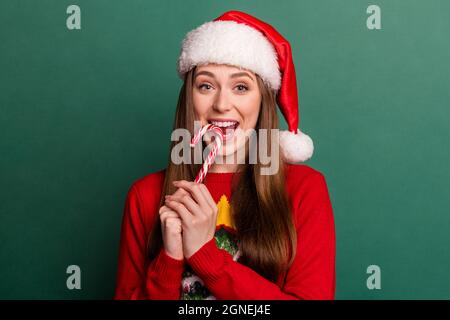 Foto di giovane donna felice sorriso positivo mangiare caramella yummy indossare santa cappello isolato su sfondo verde colore Foto Stock