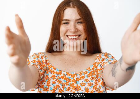 Bella ragazza rossa con un sorriso luminoso, allungando le mani per tenerti, abbracciando qualcuno, in piedi su sfondo bianco Foto Stock