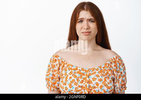 Ritratto di giovane ragazza rossa che guarda scettico, bracconda e guarda a qualcosa di strano, avendo dubbi, in piedi su sfondo bianco Foto Stock