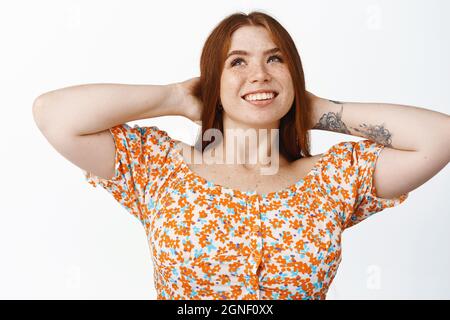 Ragazza redhead rilassata che riposa, sdraiata con le mani dietro la testa e guarda felice, riposa in vacanza, in piedi su sfondo bianco Foto Stock