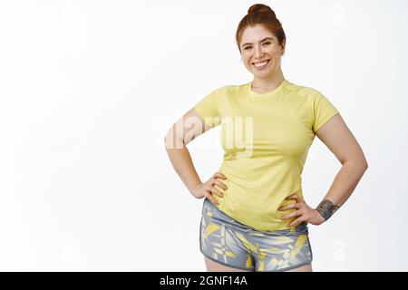 Ritratto di Happy Plus dimensione ragazza allenamento in palestra, vestito sportivo e sorridente alla macchina fotografica, in piedi su sfondo bianco Foto Stock