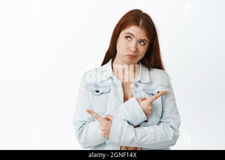 Confusa giovane donna rossa che punta lateralmente e guarda verso l'alto, pensando a cosa scegliere, scegliendo tra due varianti, sfondo bianco Foto Stock