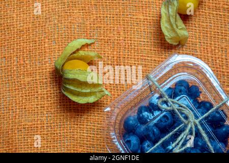 Frutti di physalis freschi e maturi e mirtilli in scatola di plastica trasparente giacciono su un burlap di colore arancione. Alimentazione sana, dieta. Vista dall'alto. Spazio di copia. Foto Stock