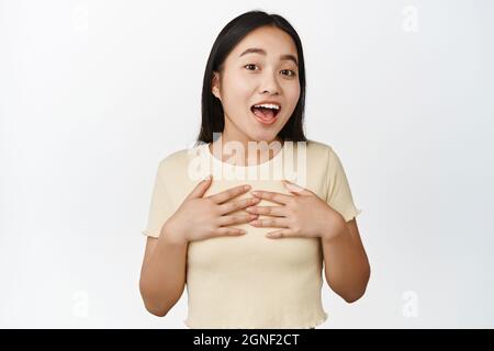 Donna asiatica entusiasta che tiene le mani sul petto, sorridendo e guardando eccitato, stupito da qualcosa, in piedi su sfondo bianco Foto Stock