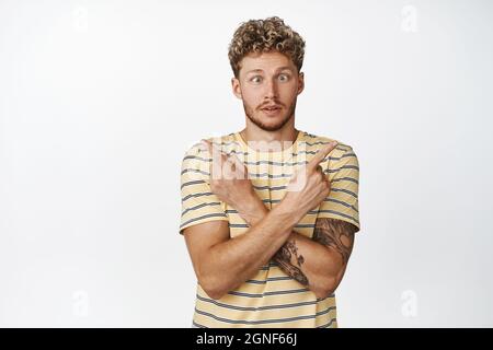 Il ragazzo curly biondo divertente punta le dita lateralmente, gli occhi squinting e mostrando la varietà di scelte in negozio, cercando di prendere una decisione, in piedi su bianco Foto Stock