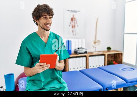 Giovane uomo ispanico che indossa uniforme fisioterapista utilizzando il touchpad presso la clinica fisioterapista Foto Stock