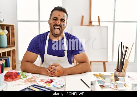 Giovane uomo ispanico con barba in studio d'arte con volto dipinto che attacca la lingua felice con espressione divertente. Concetto di emozione. Foto Stock