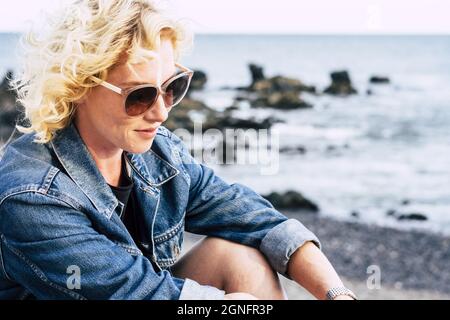Bella giovane donna in occhiali da sole sorridente e rilassante sulla spiaggia. Donna caucasica che trascorre tempo libero all'aperto. Donna elegante in denim giacca e s Foto Stock