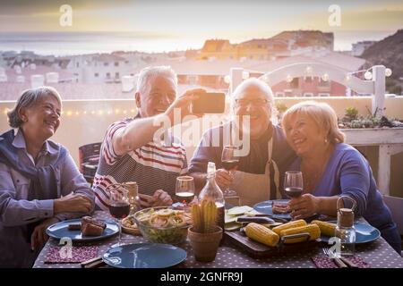 Due coppie anziane che cenano in terrazza e prendono selfie con smartphone. Gli anziani si divertono mentre cenano sul tetto. Vecchia famiglia Foto Stock