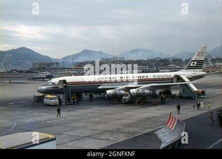 Japan Air Lines Douglas DC-8 Airliner ‘Kirishima’(JA8010) è stato reso pronto per il volo all'aeroporto di Kai Tak, Hong Kong nel 1968. Il velivolo ha lo slogan pubblicitario “Official Airline for Japan World Exposition” dipinto sulla sua fusoliera, promuovendo l’imminente “Expo '70”, la fiera mondiale tenutasi a Suita, Osaka, Giappone nel 1970. I passeggeri camminano verso l'edificio del terminal, dopo essere sbarcati dall'aereo. Un vecchio aereo Douglas DC-3 azionato da un propulsore è parcheggiato in lontananza. Foto Stock