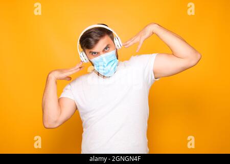 un uomo in cuffie musicali bianche si erge su uno sfondo giallo, indossando una maschera medica. Foto Stock