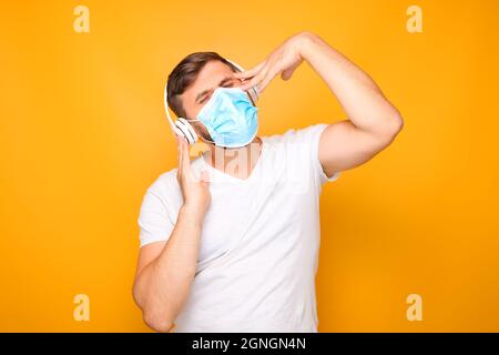 un uomo in cuffie musicali bianche si erge su uno sfondo giallo, indossando una maschera medica. Foto Stock