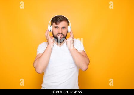 un uomo barbuto in piedi su uno sfondo giallo preme le cuffie bianche alle sue orecchie e ascolta la musica. Foto Stock