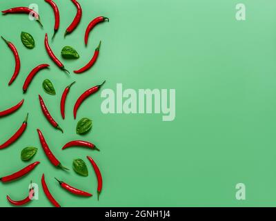 Peperoncini rossi crudi e foglie di basilico fresco su sfondo verde smeraldo. Peperoncini piccanti composizione creativa per la progettazione di alimenti. Spazio di copia. Foto Stock