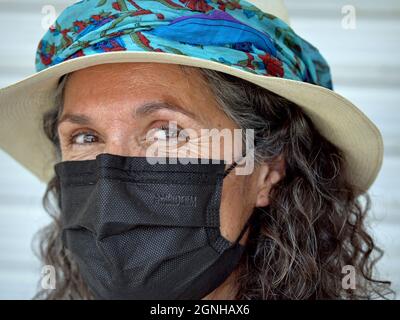 Affascinante positiva matura donna caucasica con gli occhi marroni bella indossa panama cappello con elegante blu fascia cappello e nero maschera facciale monouso. Foto Stock