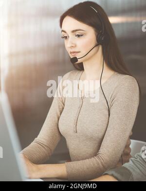 Una giovane ragazza con cuffie parla con il cliente di un'azienda, mentre si siede alla scrivania in un ufficio soleggiato. Operatori di call center al lavoro Foto Stock