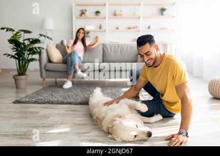 Felice ragazzo arabo stroking carino cane sul pavimento a casa, la sua ragazza rilassante con tazza di caffè su sfondo, copia spazio Foto Stock