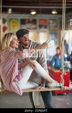 Una giovane donna sta prendendo un selfie con la sua giovane collega maschile mentre si rilassa su un swing durante una pausa in un'atmosfera rilassata in ufficio. Occupate Foto Stock