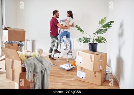 coppia caucasica innamorata divertirsi mentre si prepara a dipingere il muro bianco in appartamento, in piedi su una scala, tenendo pennelli, sorridendo, ridendo, l Foto Stock
