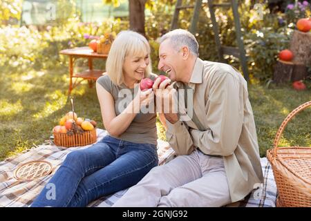 Coppia anziana positiva seduta sul campo erba e godere di mangiare mela, chiacchierando e ridendo, riposando all'aperto in giardino Foto Stock