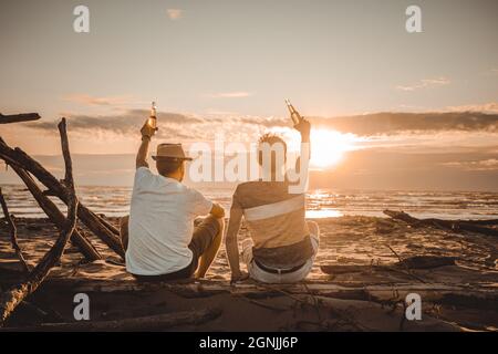 Due giovani amici trascorrono il tempo insieme seduti sulla spiaggia, bevendo birra e tostando in vacanza al tramonto estivo al crepuscolo. Concetto di amicizia Foto Stock