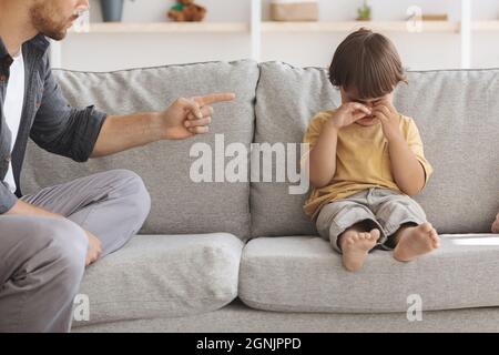 Concetto di abuso di minori. Ragazzino infelice seduto sul divano e piangendo, padre arrabbiato irriconoscibile che spaventa il bambino sconvolto Foto Stock