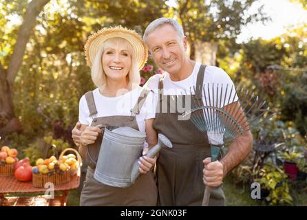 Sposi anziani graziosi che tengono attrezzi di giardinaggio, che lavorano in giardino al giorno di sole, donna con annaffiatura lattina, uomo con rastrello Foto Stock