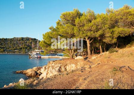 Podvrske, Murter, Croazia - 26 agosto 2021: Spiaggia rocciosa, pini e barca ormeggiata dal molo nel tramonto estivo Foto Stock